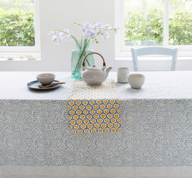 Kitchen table covered with a table cloth and laid with tea cups, a tea pot and a vase of flowers